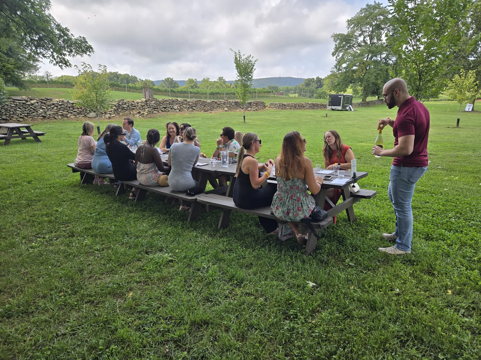 A group seated at a long picnic table on the grass with a host standing beside them and vineyard rows behind